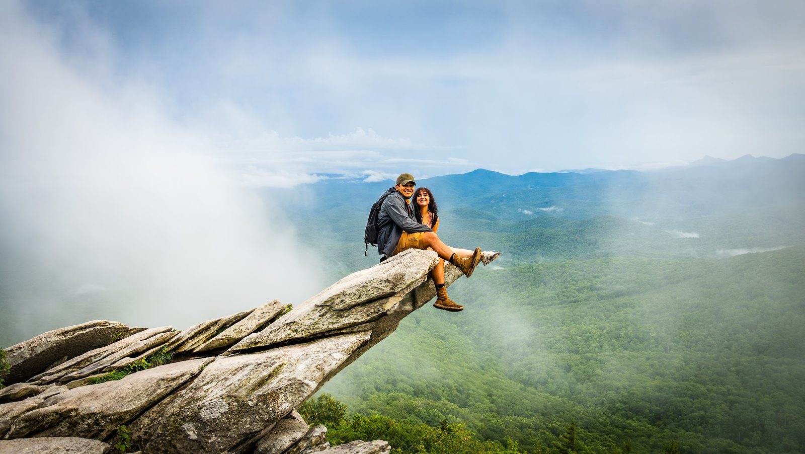 Couple sitting on cliff edge overlooking misty mountains and forest