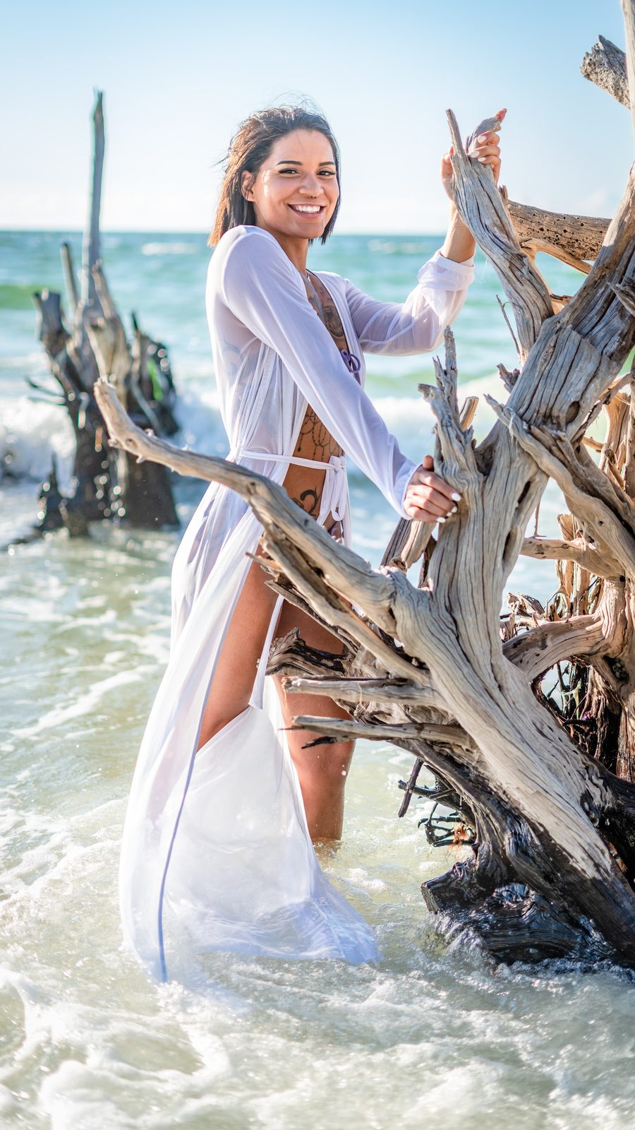 Woman in white cover-up posing with driftwood at ocean shore