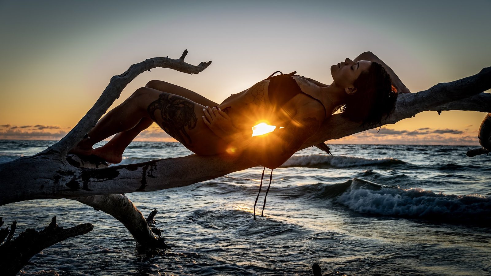 Woman posing on driftwood at sunset beach silhouette