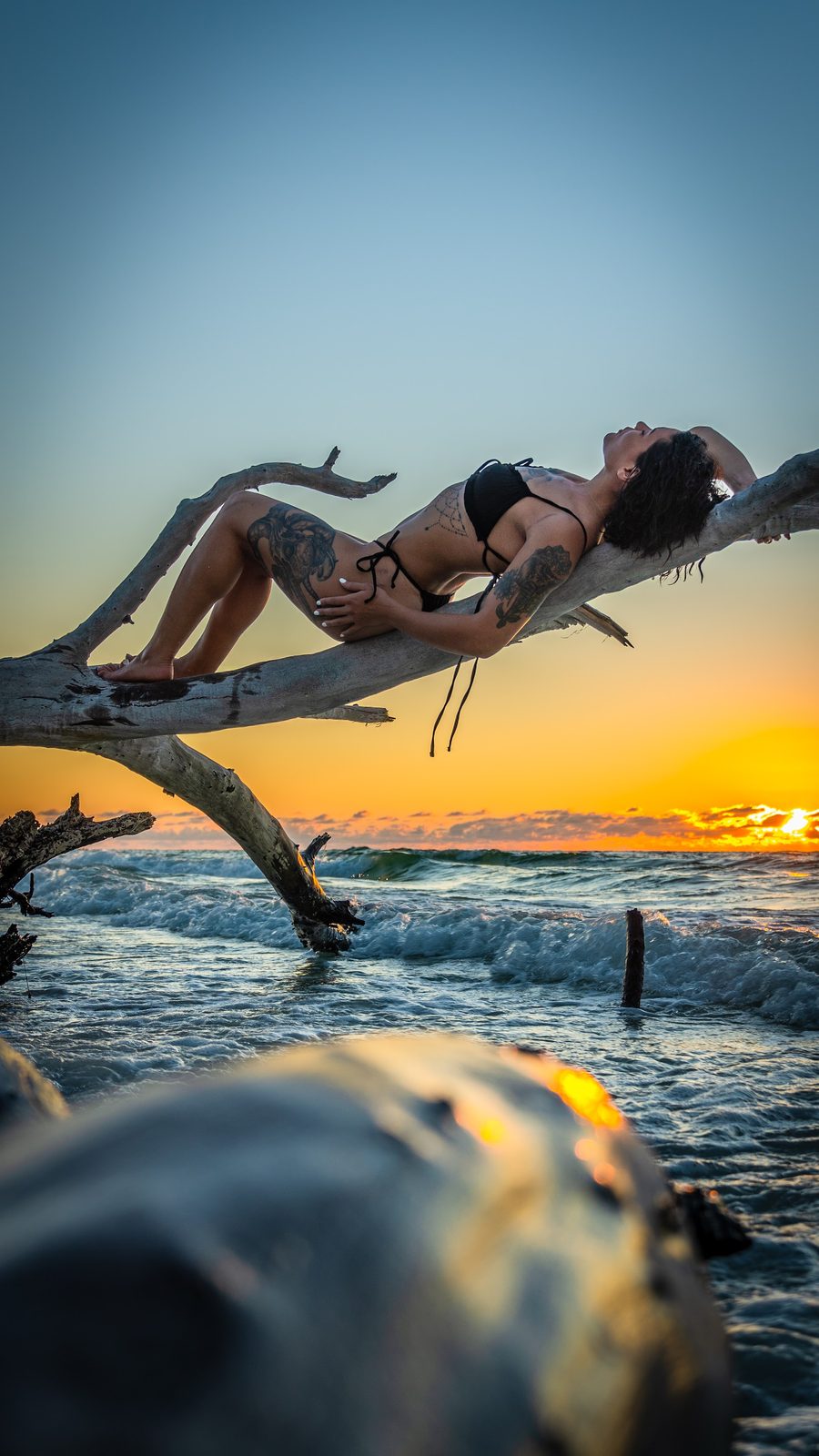 Woman in bikini posing on driftwood at sunset beach